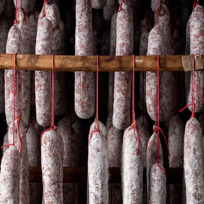 photographe artisanat marseille Photo artisan Marseille : alignement de saucissons artisanaux suspendus par des fils rouges dans un séchoir traditionnel.