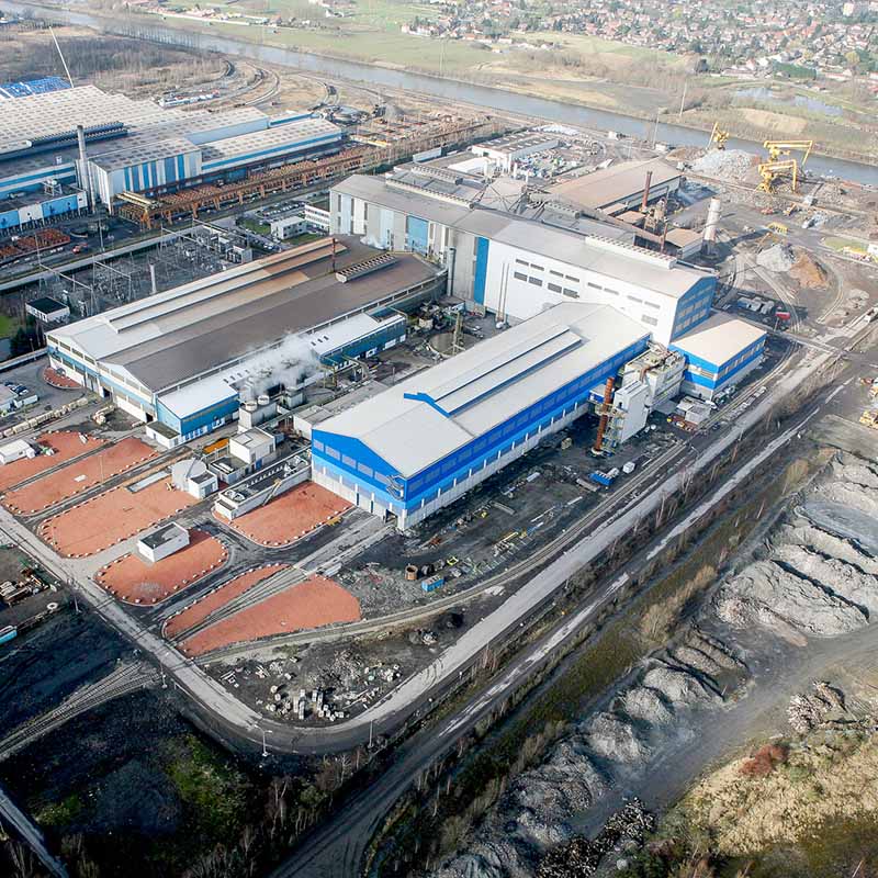 Photographie aérienne par drone d'un vaste complexe industriel avec hangars bleus, zones de stockage et canal en arrière-plan.