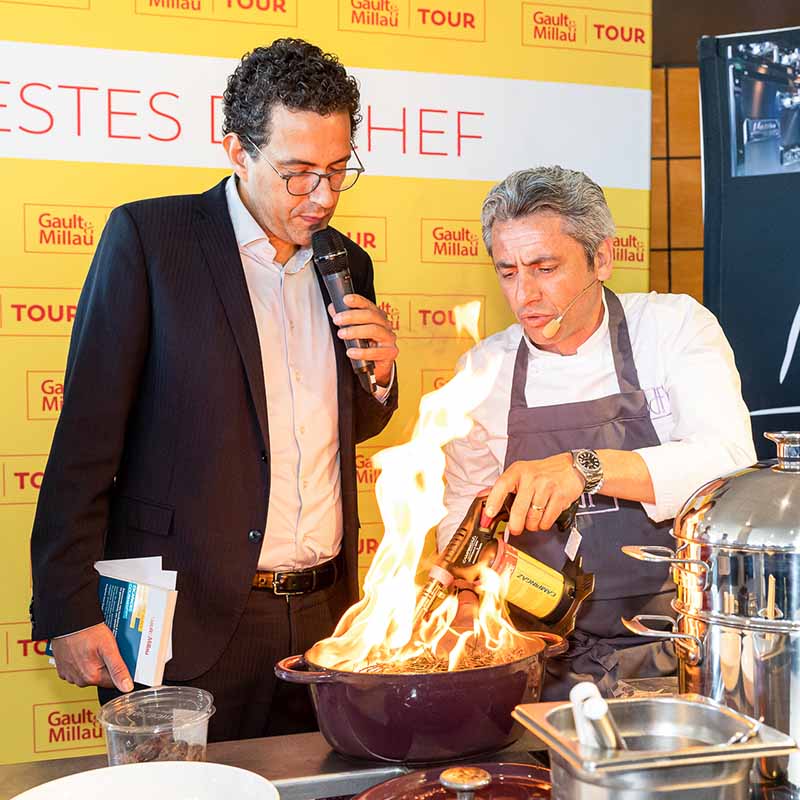 Photo événementielle Marseille : un chef cuisinier en action avec un chalumeau et des flammes lors du Gault & Millau Tour.