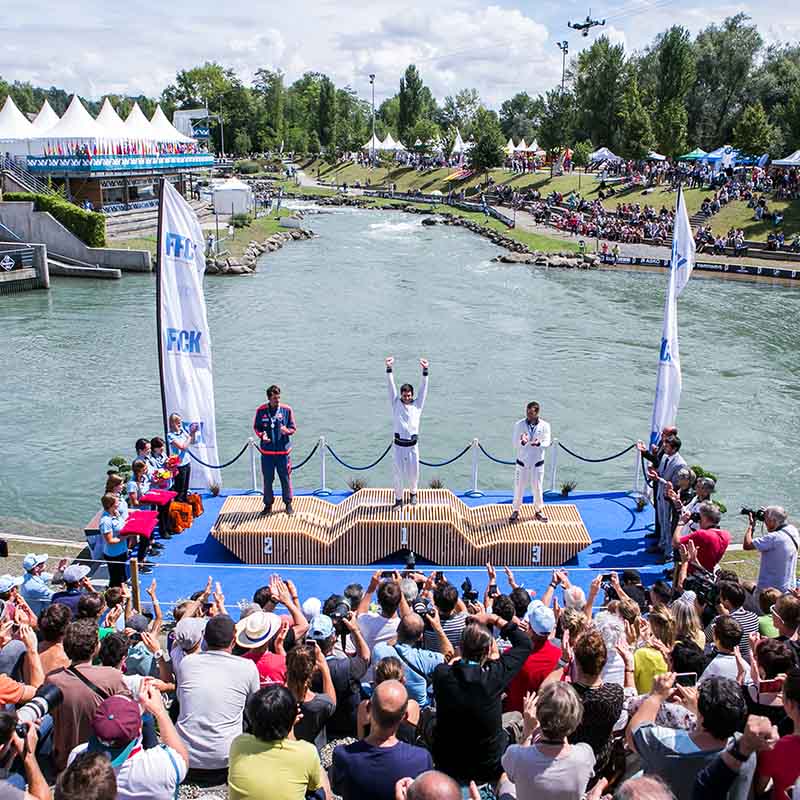 Photo événementielle Marseille : podium d'une compétition de canoë-kayak avec athlètes, public nombreux et rivière en fond.
