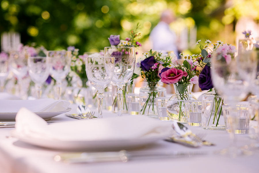 Gros plan sur une décoration de table de mariage avec des fleurs naturelles violettes et roses dans des vases en verre, vaisselle blanche et verres ciselés en extérieur à Marseille.