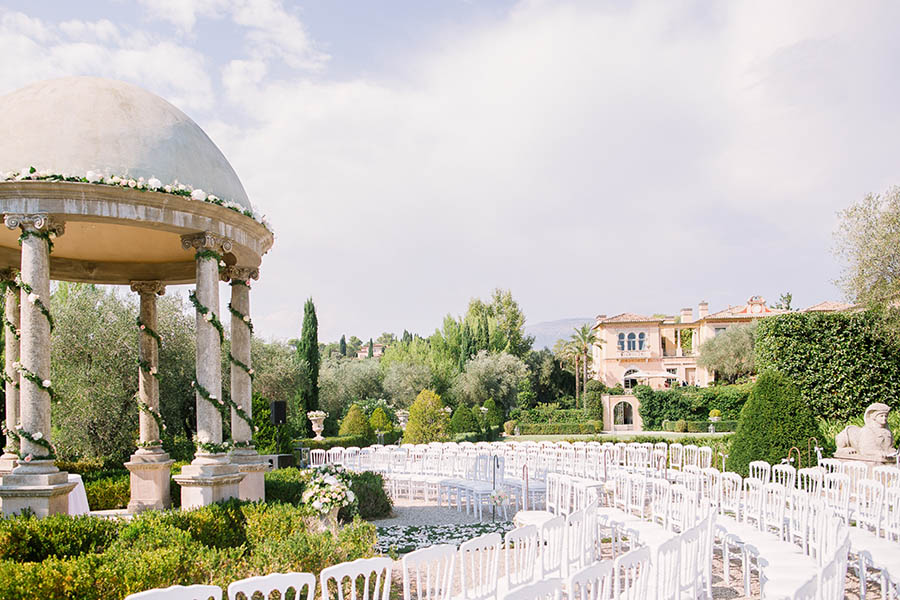 Espace de cérémonie de mariage en extérieur avec un kiosque en pierre décoré de fleurs, des rangées de chaises blanches et une bastide provençale en arrière-plan à Marseille.