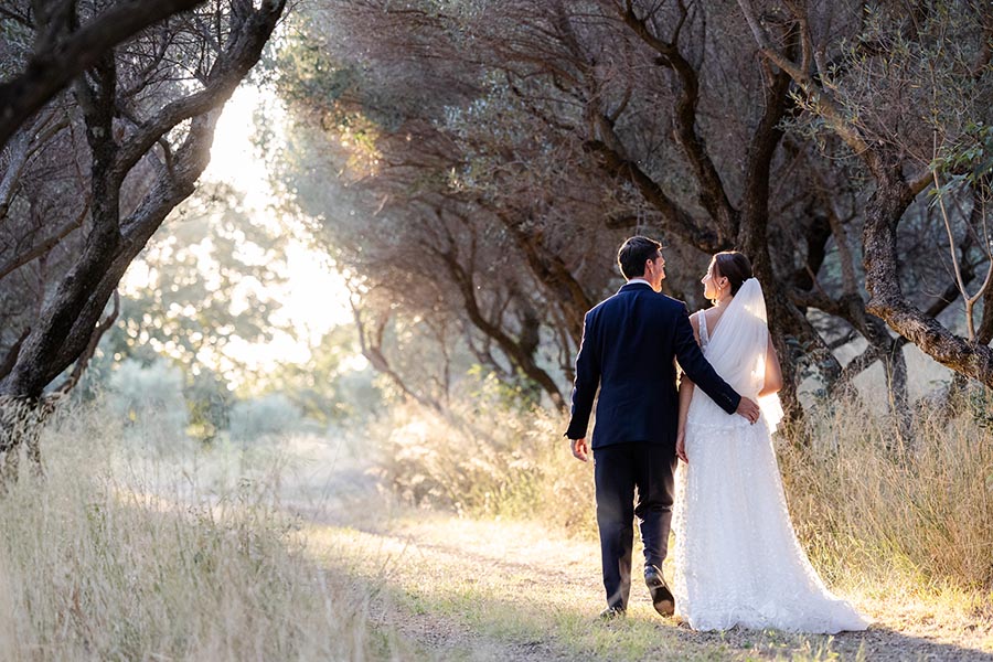 Couple de mariés marchant main dans la main dans une oliveraie à Marseille lors d'une séance photo de mariage au coucher du soleil.