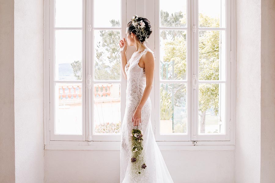 portrait mariee robe dentelle fenetre marseille Profil d'une mariée en robe de dentelle blanche posant devant une grande fenêtre lumineuse lors de ses préparatifs à Marseille.